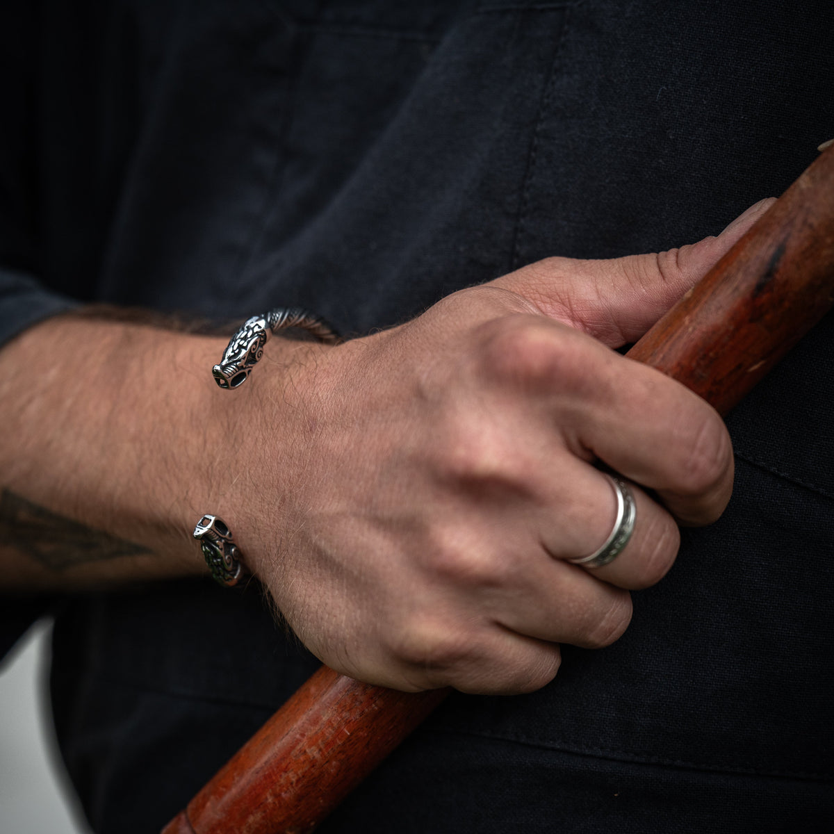 Close-up of the Fenrir Viking bracelet with detailed wolf heads, worn on a wrist while holding a wooden handle, by Ravenritual.