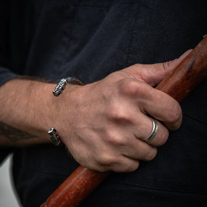 Close-up of the Fenrir Viking bracelet with detailed wolf heads, worn on a wrist while holding a wooden handle, by Ravenritual.