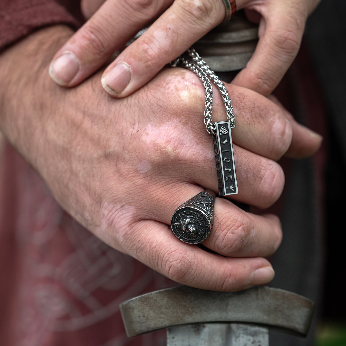 Close-up of the Fenrir Viking Ring with an engraved wolf design, worn on a hand holding a sword with a silver runic necklace draped over the fingers.