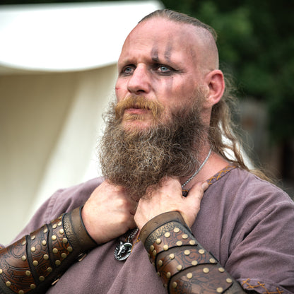 Bearded Viking-inspired warrior wearing a pendant necklace, brown tunic and leather wrist guards in an outdoor setting.