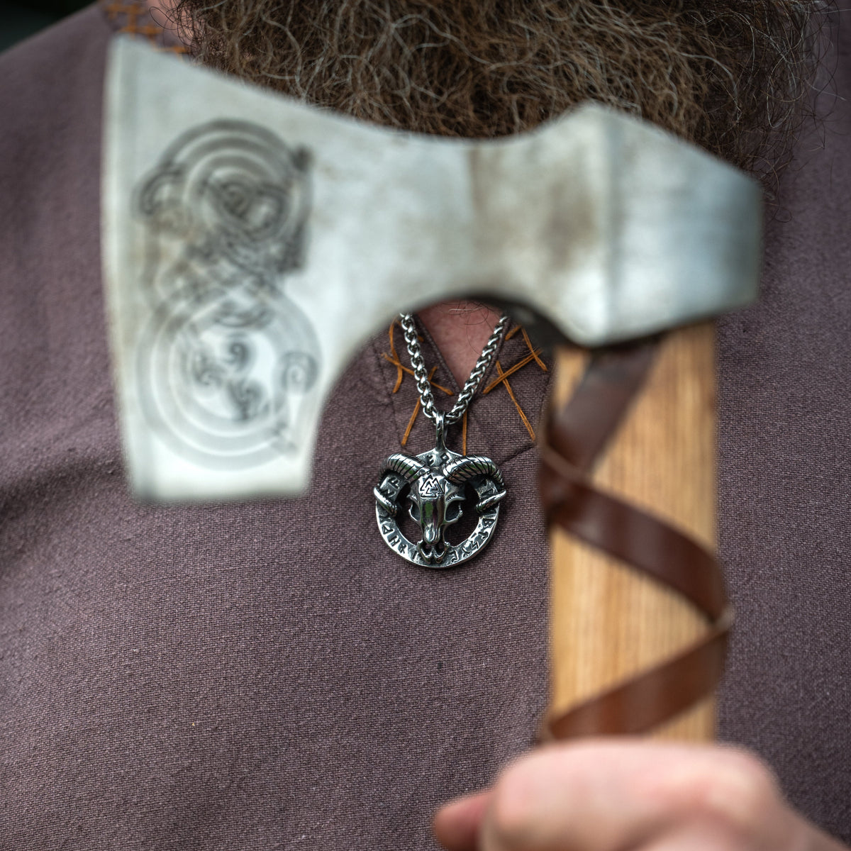 Heidrun Necklace with a silver-tone Norse goat pendant worn over a brown Viking tunic beside a traditional-style axe.
