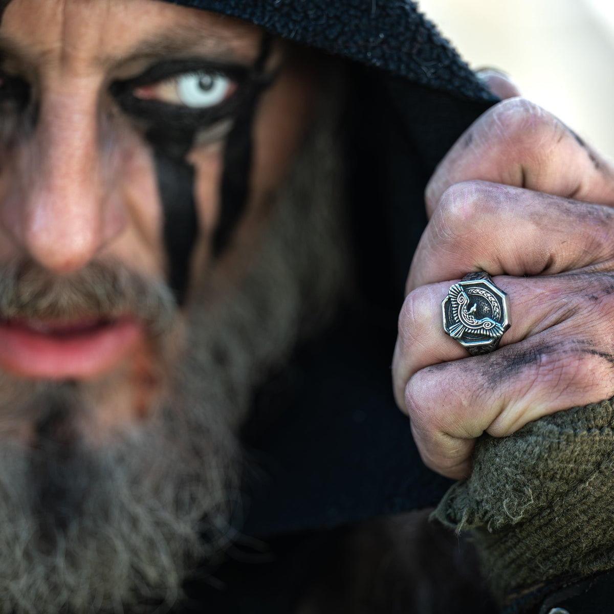 Hooded Viking-inspired warrior with black face paint displaying an engraved silver-tone Raven Ring near his face.