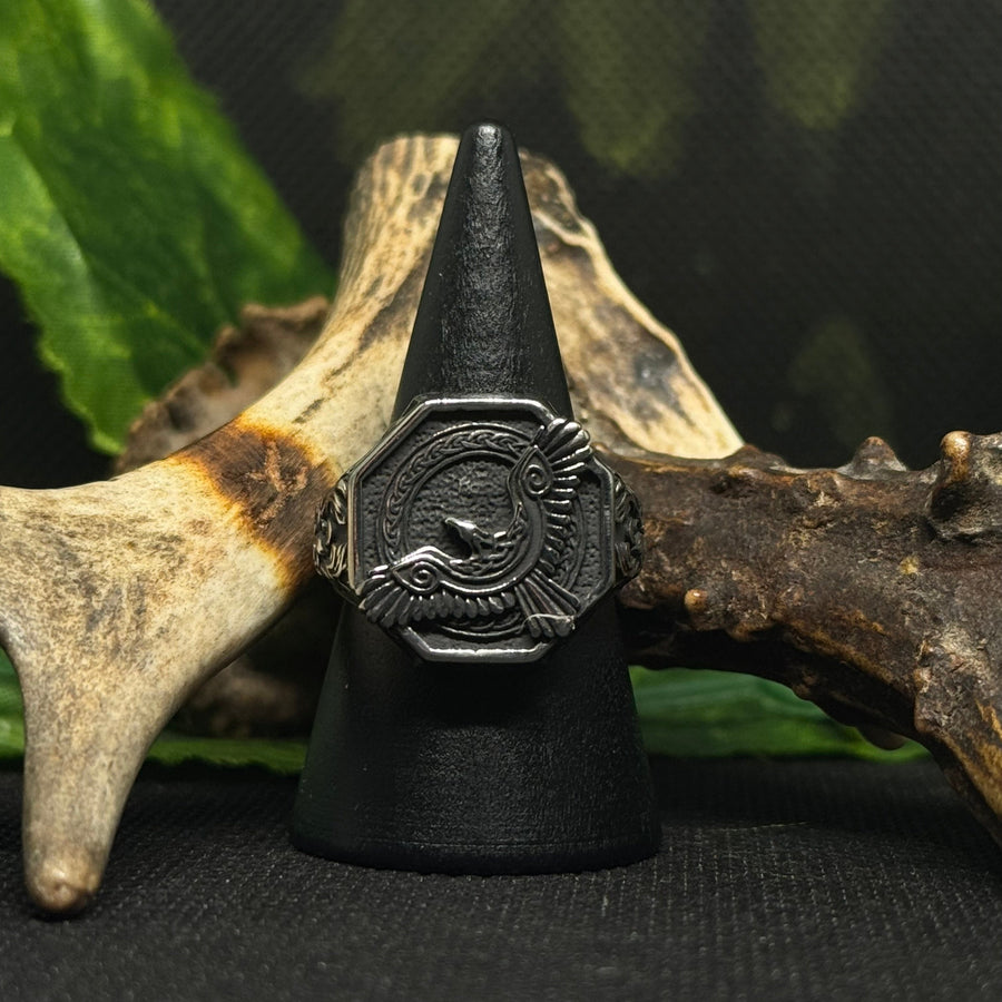 Close-up of the Raven Ring featuring an engraved Norse raven design, displayed on a black stand with antlers and greenery.