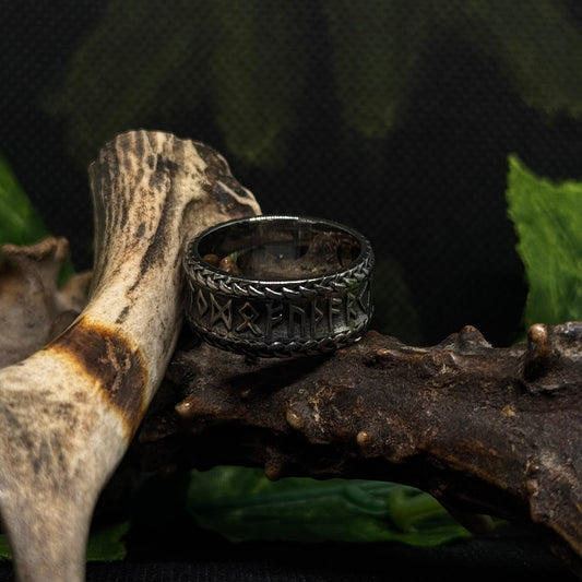 Close-up of the Ravenritual Rune Ring with engraved Norse runic symbols, displayed on rustic wood beside antlers and greenery.