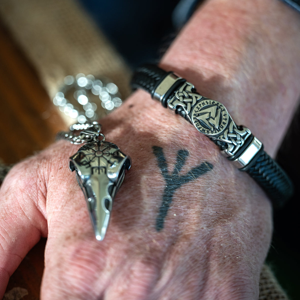 Close-up of the Valknut Bracelet with engraved Norse knotwork and braided black cord, worn on a wrist beside other Viking jewelry.
