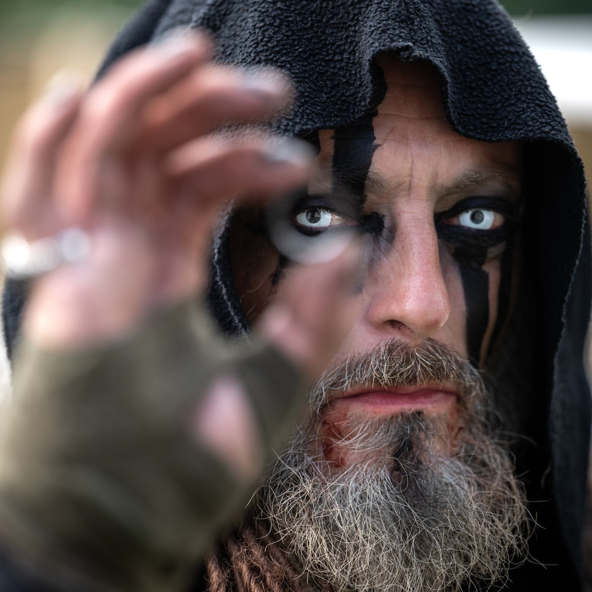 Close-up portrait of a hooded Viking warrior with black face paint, pale eyes and a grey beard, reaching toward the camera.