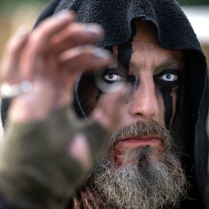 Close-up portrait of a hooded Viking warrior with black face paint, pale eyes and a grey beard, reaching toward the camera.